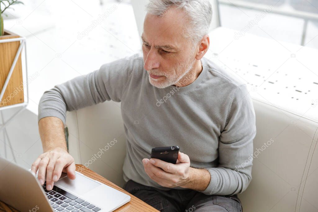 Photo of focused gray-haired man working with laptop and cellphone while sitting in cafe indoors
