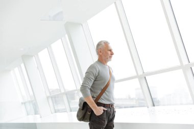 Yakışıklı, gri saçlı bir adamın fotoğrafı. Poz verip ellerini ceplerinde tutarken pencerelerin yanında duruyor.