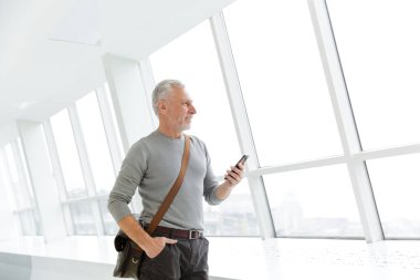 İçerideki pencerelerin yanında duran, cep telefonunda yazan, çantalı, yakışıklı, gri saçlı adamın fotoğrafı.