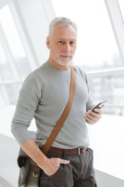İçerideki pencerelerin yanında duran, cep telefonunda yazan, çantalı, yakışıklı, gri saçlı adamın fotoğrafı.