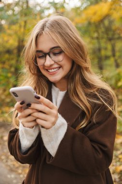 Güz parkında yürürken cep telefonu kullanan güzel ve neşeli bir kadın.