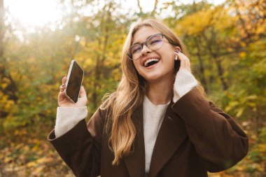 Güz parkında palto giyen, kablosuz kulaklık takan, cep telefonu kullanan, müzik dinleyen güzel bir genç kadın.