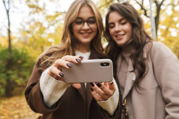 2人の陽気な若いかわいい女の子がコートを着て一緒に秋の森を歩いて 携帯電話を見て — ストック写真