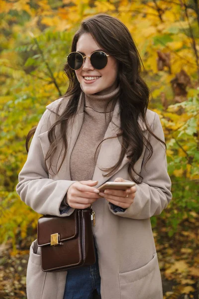 美丽的年轻而快乐的女人穿着外套走在秋天的公园里 用手机 — 图库照片