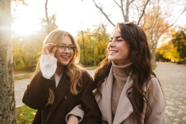 Sonbahar parkında birlikte yürüyen ceketli iki neşeli genç kız.