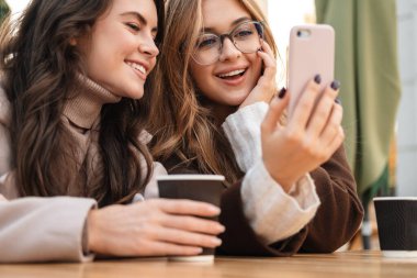 İki neşeli çekici bayan arkadaş kafede oturup kahve içerken cep telefonlarına bakıyorlar.