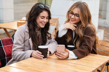 İki neşeli çekici bayan arkadaş kafede oturup kahve içerken cep telefonlarına bakıyorlar.