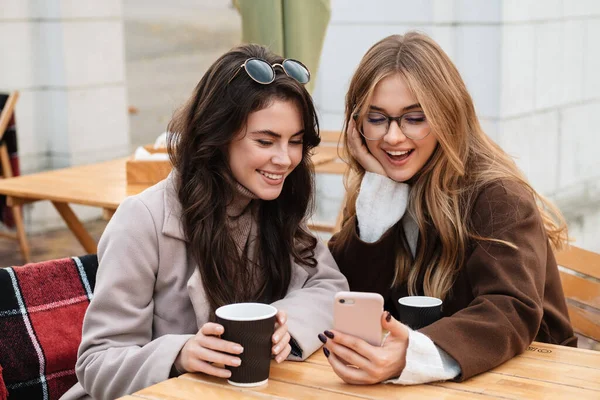 2人の明るい魅力的な女性の友人が屋外のカフェに座って コーヒーを飲みながら携帯電話を見て — ストック写真