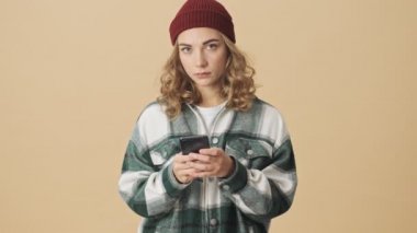 Örgü şapkalı, gömlekli, akıllı telefon kullanan ve bej arka planda mutlu olan güzel bir kadın.