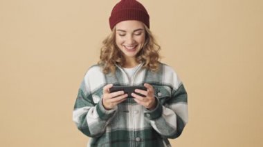 Örgü şapkalı ve gömlekli neşeli güzel kadın bej arka planda akıllı telefonla oynuyor.