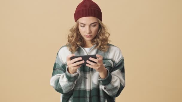 ニット帽とシャツを身に着けているかなり女性は ベージュの背景にスマートフォンを使用している間に怖がっている — ストック動画