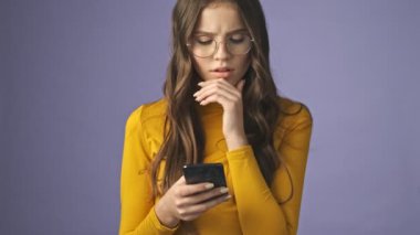 Düşünceli, ciddi bir genç kız, atölyedeki mor arka planda tek başına duran akıllı telefonunu kullanıyor.