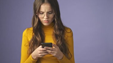 Düşünceli, ciddi bir genç kız telefonuyla sohbet ediyor. Stüdyonun mor arka planında tek başına duruyor.