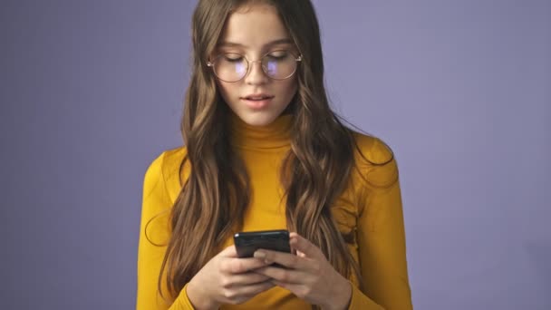 一个忙碌的少女正在用她的智能手机聊天 一边认出一个人 一边向他挥手 一边孤零零地站在工作室的紫色背景下 — 图库视频影像