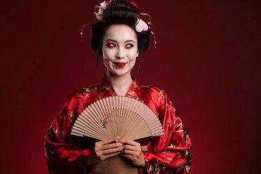 Geleneksel Japon kimonosu giymiş genç bir geyşa resmi. Kırmızı arka planda izole edilmiş ahşap el yelpaze tutuyor.
