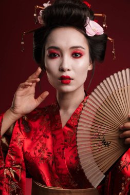 Geleneksel Japon kimonosu giymiş genç bir geyşa resmi. Kırmızı arka planda izole edilmiş ahşap el yelpaze tutuyor.