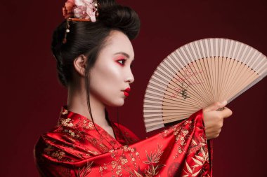 Geleneksel Japon kimonosu giymiş genç bir geyşa resmi. Kırmızı arka planda izole edilmiş ahşap el yelpaze tutuyor.