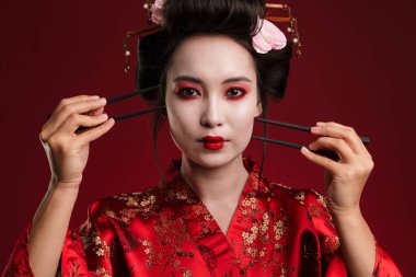 Geleneksel Japon kimonosu giymiş güzel bir geyşa kadın resmi kırmızı arka planda izole edilmiş çubuklar tutuyor.