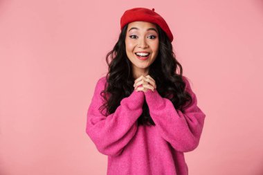 Uzun siyah saçlı, neşeli Asyalı bir kızın görüntüsü. Bere takmış pembe arka planda izole edilmiş kameraya gülüyor.