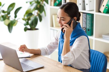 Ofiste çalışan çekici gülümseyen Asyalı iş kadını dizüstü bilgisayarla çalışırken cep telefonuyla konuşuyor.