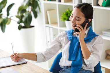 Ofiste dizüstü bilgisayarla çalışırken cep telefonuyla konuşan Asyalı genç bir sekreter resmi