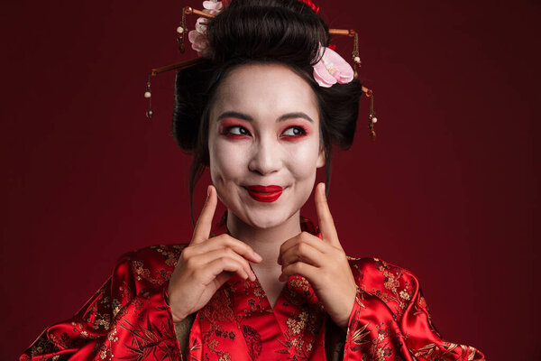 Image of beautiful young geisha woman in traditional japanese kimono smiling isolated over red background