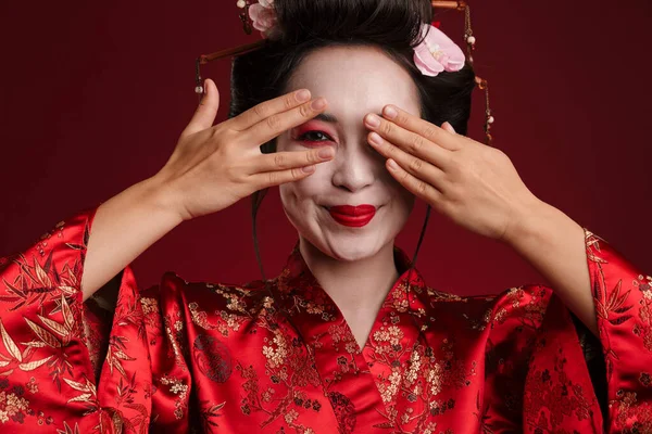 穿着日本传统和服的年轻漂亮的艺妓的照片 她的眼睛被隔离在红色背景下的双手遮住 — 图库照片