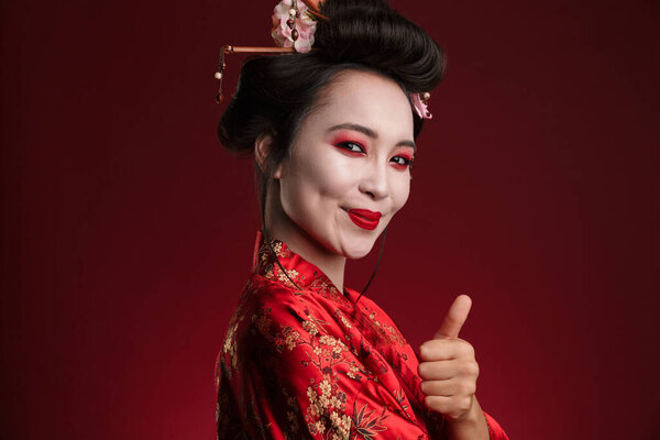 Image of gorgeous geisha woman in traditional japanese kimono showing thumb up isolated over red background