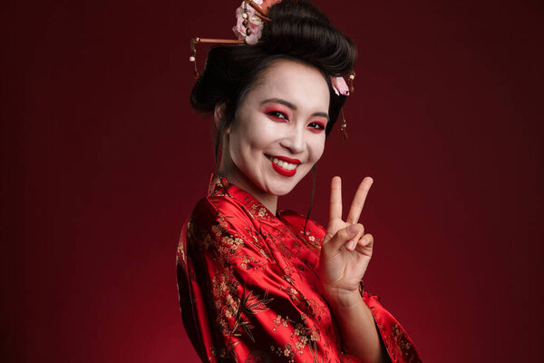 Image of gorgeous geisha woman in traditional japanese kimono showing peace sign isolated over red background