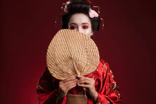 照片中身穿日本传统和服的年轻艺妓 手持红色背景的木制手扇 — 图库照片