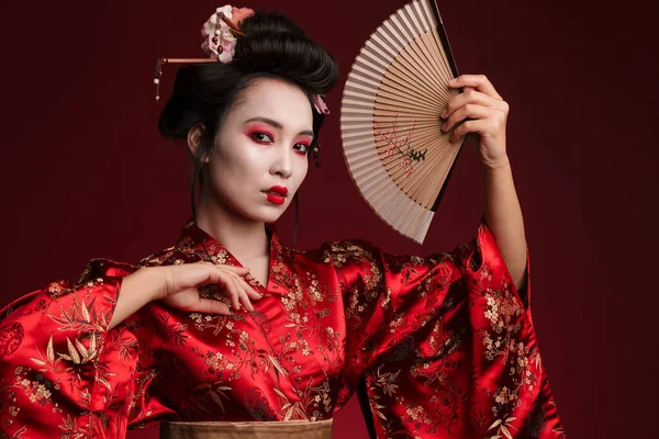 照片中身穿日本传统和服的年轻艺妓 手持红色背景的木制手扇 — 图库照片