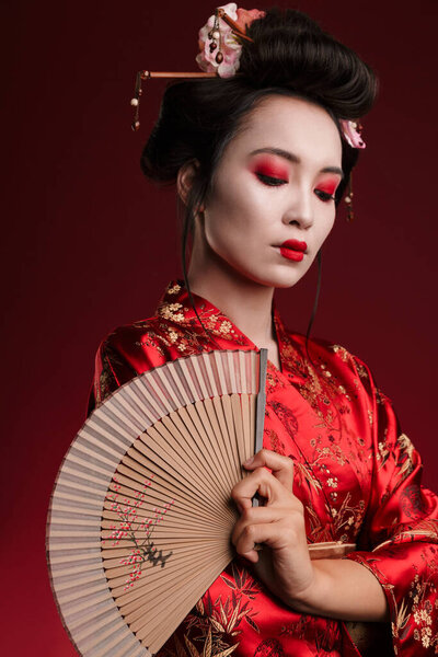 Image of young geisha woman in traditional japanese kimono holding wooden hand fan isolated over red background