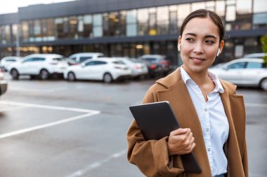 Sırıtkan Asyalı genç iş kadını. Şehirde palto giyiyor, dizüstü bilgisayar taşıyor.