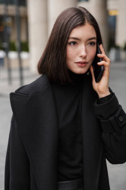 Sonbahar ceketi giyen çekici, genç esmer kadın bir caddede duruyor, cep telefonu kullanıyor.