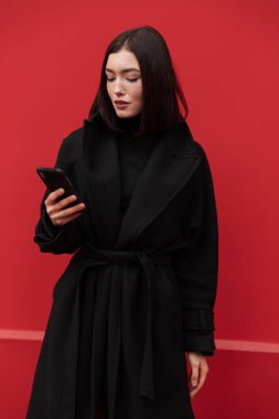 Sonbahar ceketi giyen güzel esmer kadın pembe arka planda tek başına duruyor, elinde cep telefonu var.