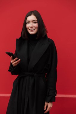 Sonbahar ceketi giyen güzel, gülümseyen esmer kadın pembe arka planda tek başına duruyor, elinde cep telefonu var.
