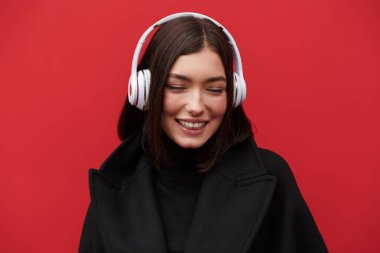 Güzel, gülümseyen, sonbahar ceketi giyen, pembe arka planda izole edilmiş, kablosuz kulaklıkla müzik dinleyen esmer bir kadın.