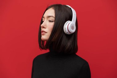 Sonbahar ceketi giyen güzel, genç, esmer bir kadın pembe arka planda dikilip kablosuz kulaklıkla müzik dinliyor.