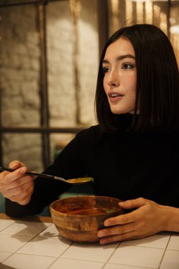 Güzel, genç esmer kadın, kafeterya masasında oturuyor, çorba içiyor.