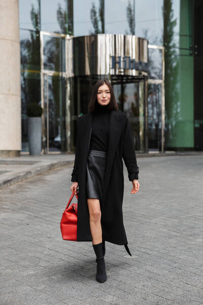 Attractive young brunette woman wearing autumn coat walking in a street, carrying travel bag