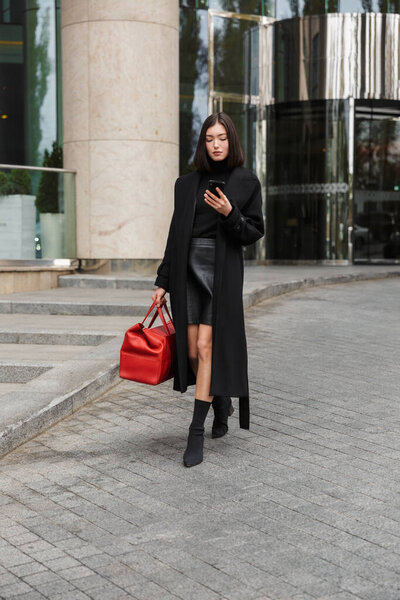 Attractive young brunette woman wearing autumn coat walking in a street, using mobile phone, carrying travel bag