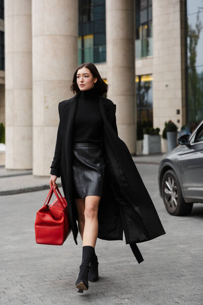 Attractive young brunette woman wearing autumn coat walking in a street, carrying travel bag