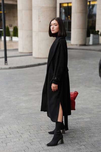 Attractive young brunette woman wearing autumn coat standing in a street, carrying travel bag
