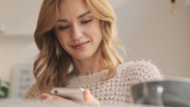 Güzel gülümseyen genç bir kadın, güneşli bir günde rahat bir kafede akıllı telefonunu tutarken kameraya bakıyor.