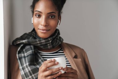 Güzel, gülümseyen, sonbahar kıyafeti giyen, duvara yaslanmış, kablosuz kulaklıkla müzik dinleyen, kahve içen Afrikalı bir kadın.