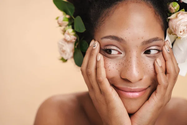 Portrait of young half-naked freckled african american woman with flowers in her hair isolated over beige background