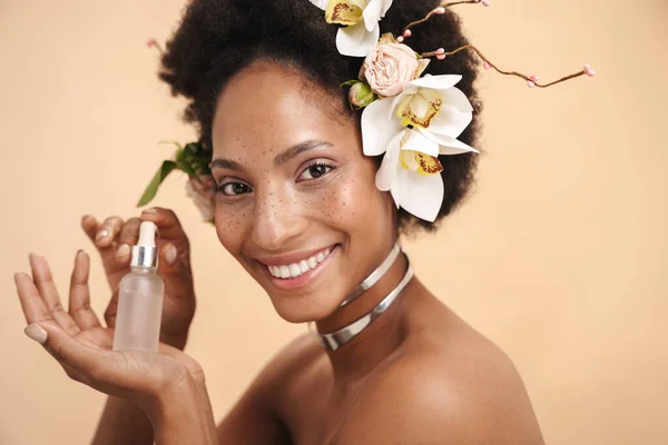 Portrait of young freckled african american woman holding facial serum cream bottle isolated over beige background