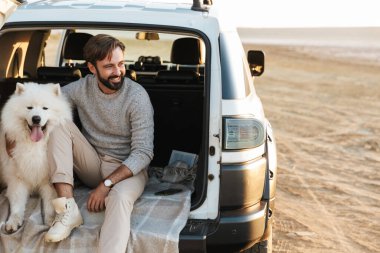 Yakışıklı genç sakallı adam arabasının arkasında oturuyor, sahilde köpekle oynuyor.