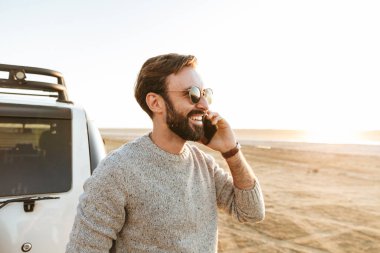 Gülümseyen yakışıklı genç adam, güneşli sahilde arabanın yanında dururken cep telefonu kullanıyor.