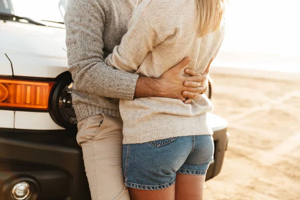 日当たりの良いビーチで車にもたれながら愛を抱いている若いカップルの作物のイメージ — ストック写真